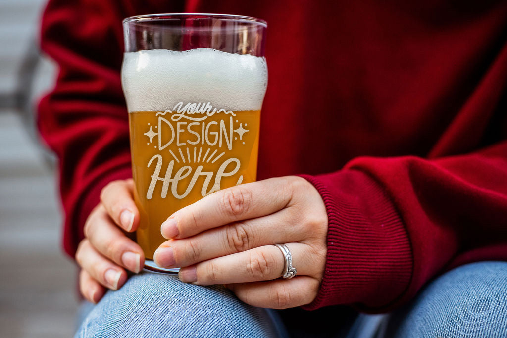 A zoomed in photo of a person in a red sweater, holding a beer glass on their knee. The glass is filled with beer and laser engraved with "Your Design Here"