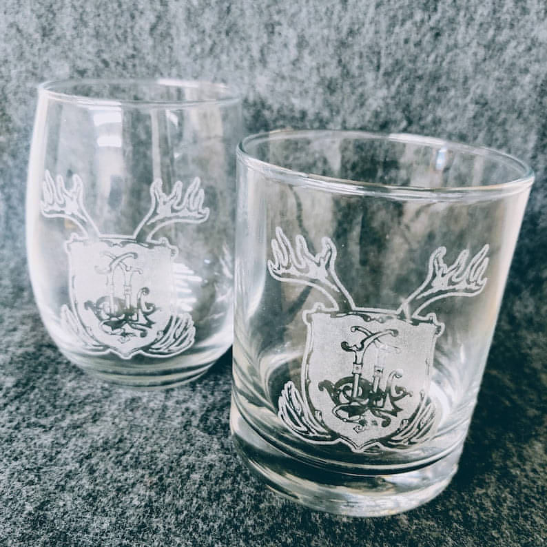 A whiskey glass and a stemless wine glass each engraved with a coat of arms