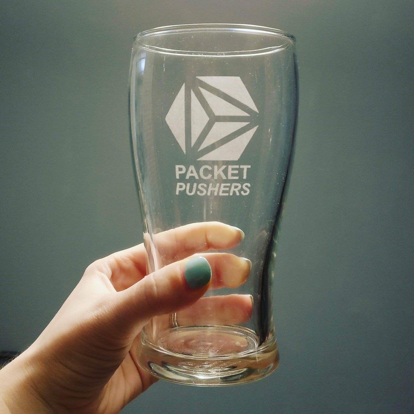A pint glass being held up, engraved with the logo for Packet Pushers